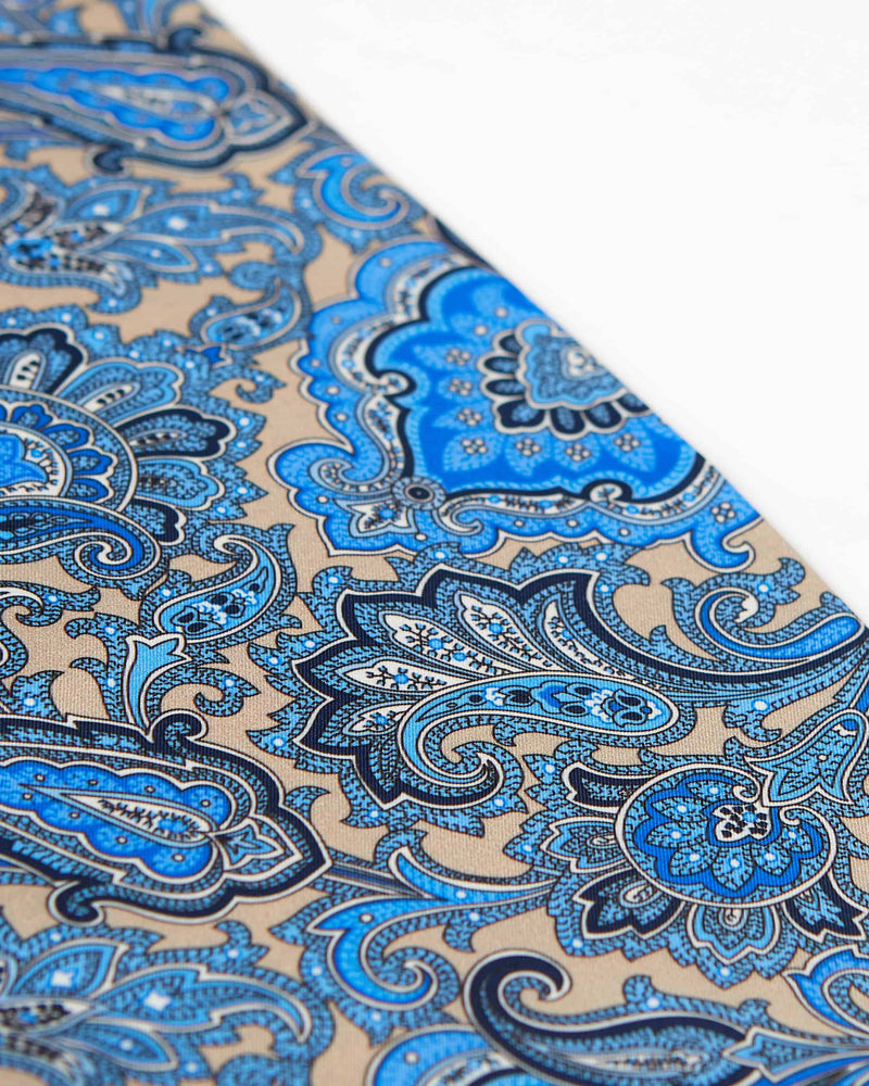 Closer, diagonal angled view of the blue paisley patterns on a cream ground of 'The Piccadilly' wool-silk scarf.
