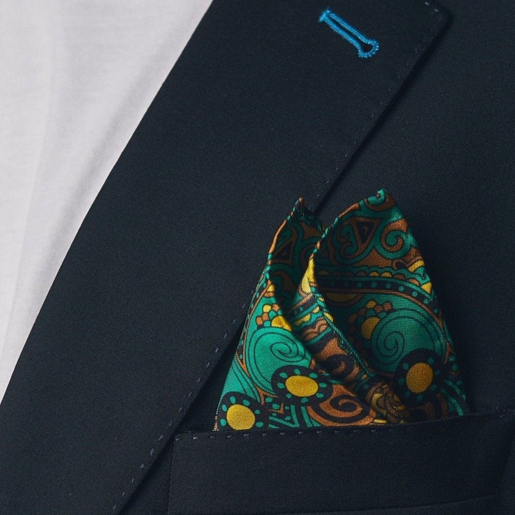 Silk square handkerchief with retro-inspired pattern on a sapphire-green background. Presented in a ‘cone’ and placed in a jacket breast pocket.