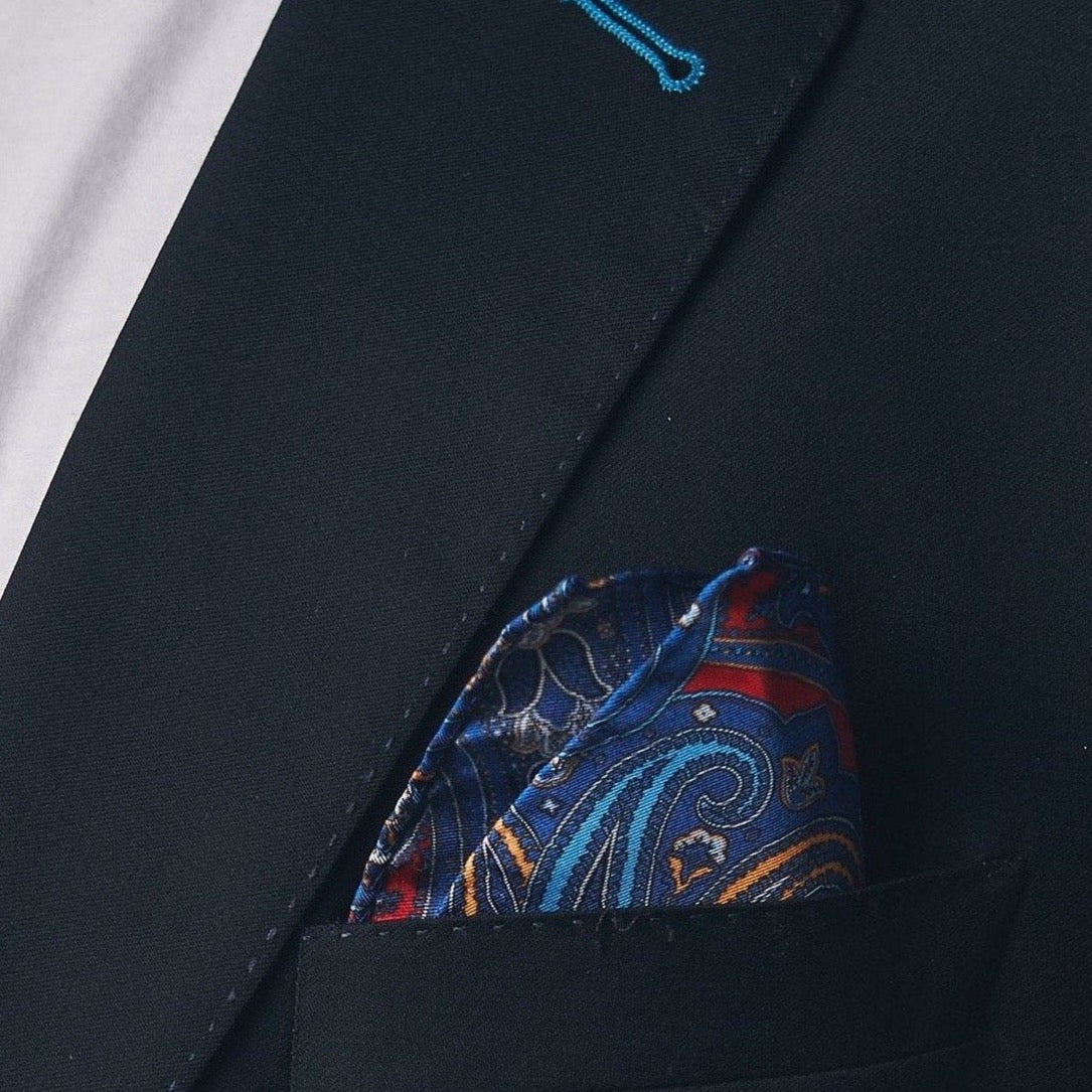 Silk square handkerchief presented in a ‘cone’ and placed in a suit jacket pocket, showing the showing the blue paisley patterns and surrounding floral patterns.