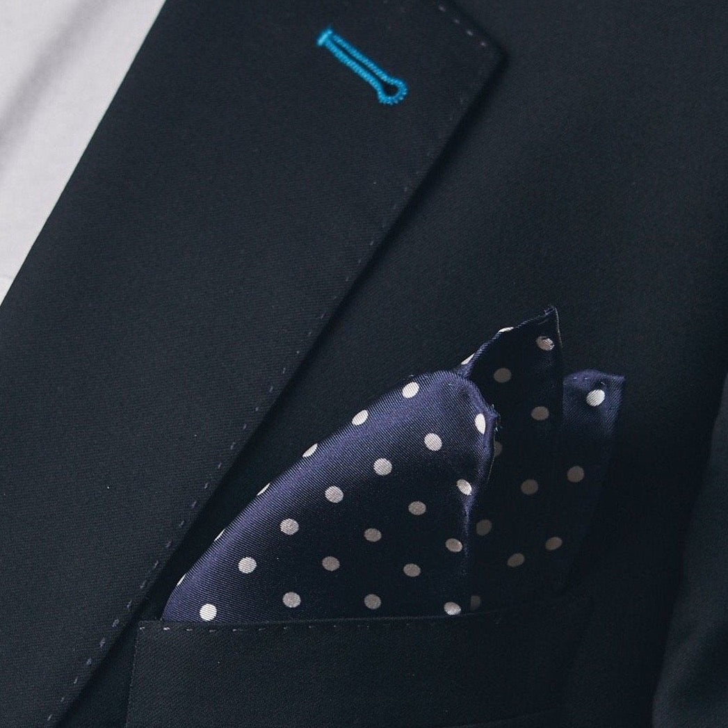Silk square handkerchief presented in a ‘cone’ and placed in a suit jacket pocket, showing the navy-blue fabric complemented by small white dots.